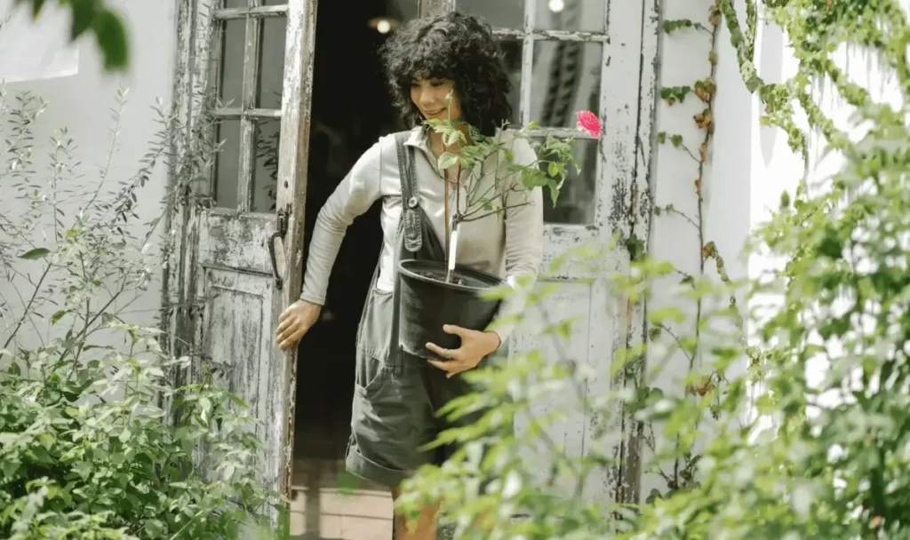 A woman with a flowering plant in a bucket enters her garden.