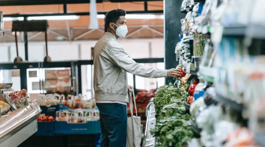 Man with a reusable bag, selecting fresh vegetables in a store for sustainable shopping.